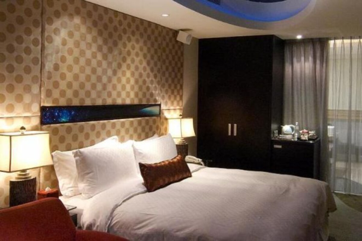 A hotel room with a large bed, a nightstand with a lamp, a wardrobe, and a view of the outside through a window.