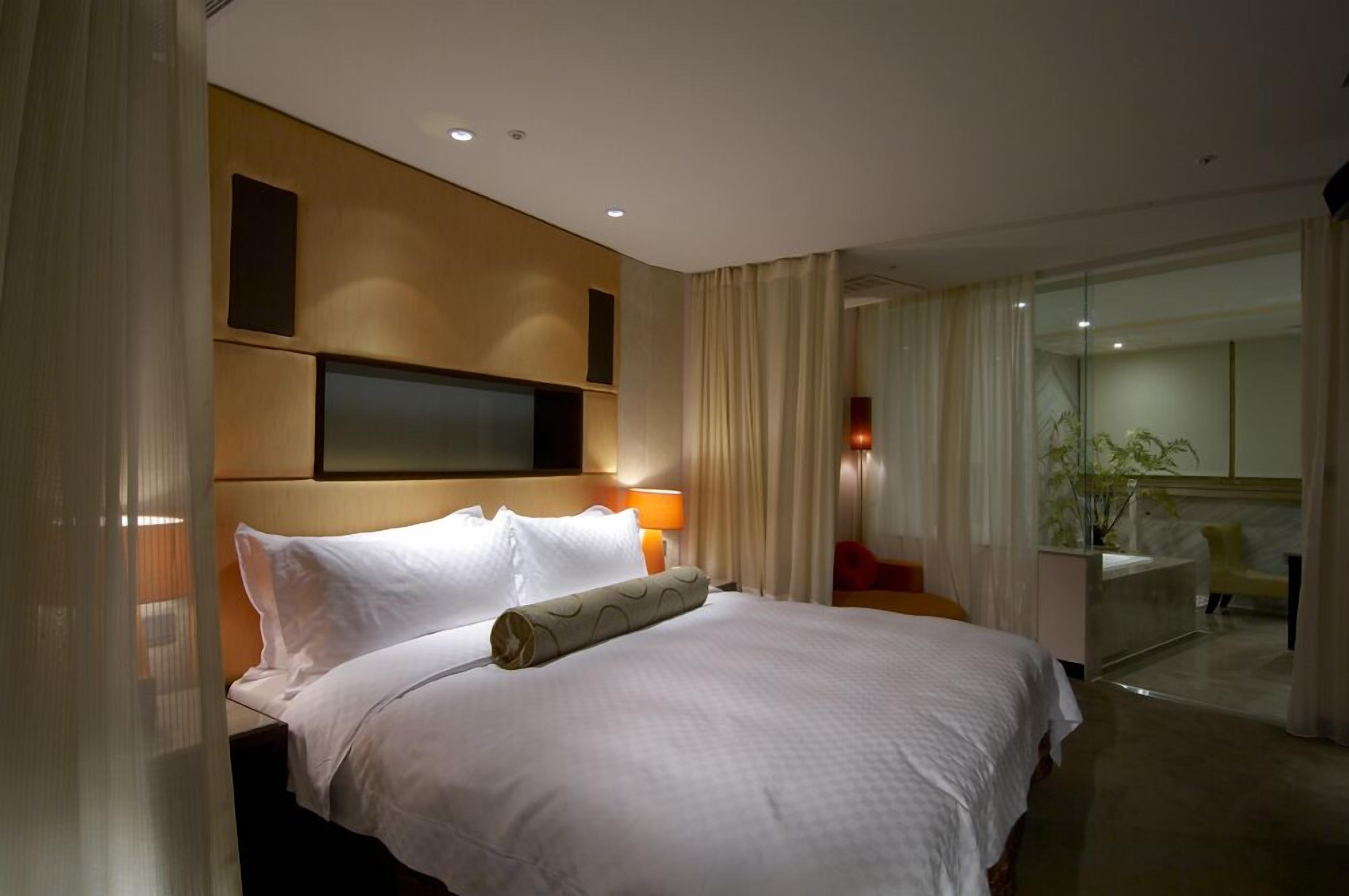 A hotel room with a large bed, a flat-screen TV mounted on the wall, a bedside table with a lamp, and a view of a balcony with a chair.