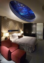 Een hotelkamer met een bed, een rode fauteuil, een nachtkastje, een lamp, een tv aan de muur en een plafond met een sterrenhemelmotief.