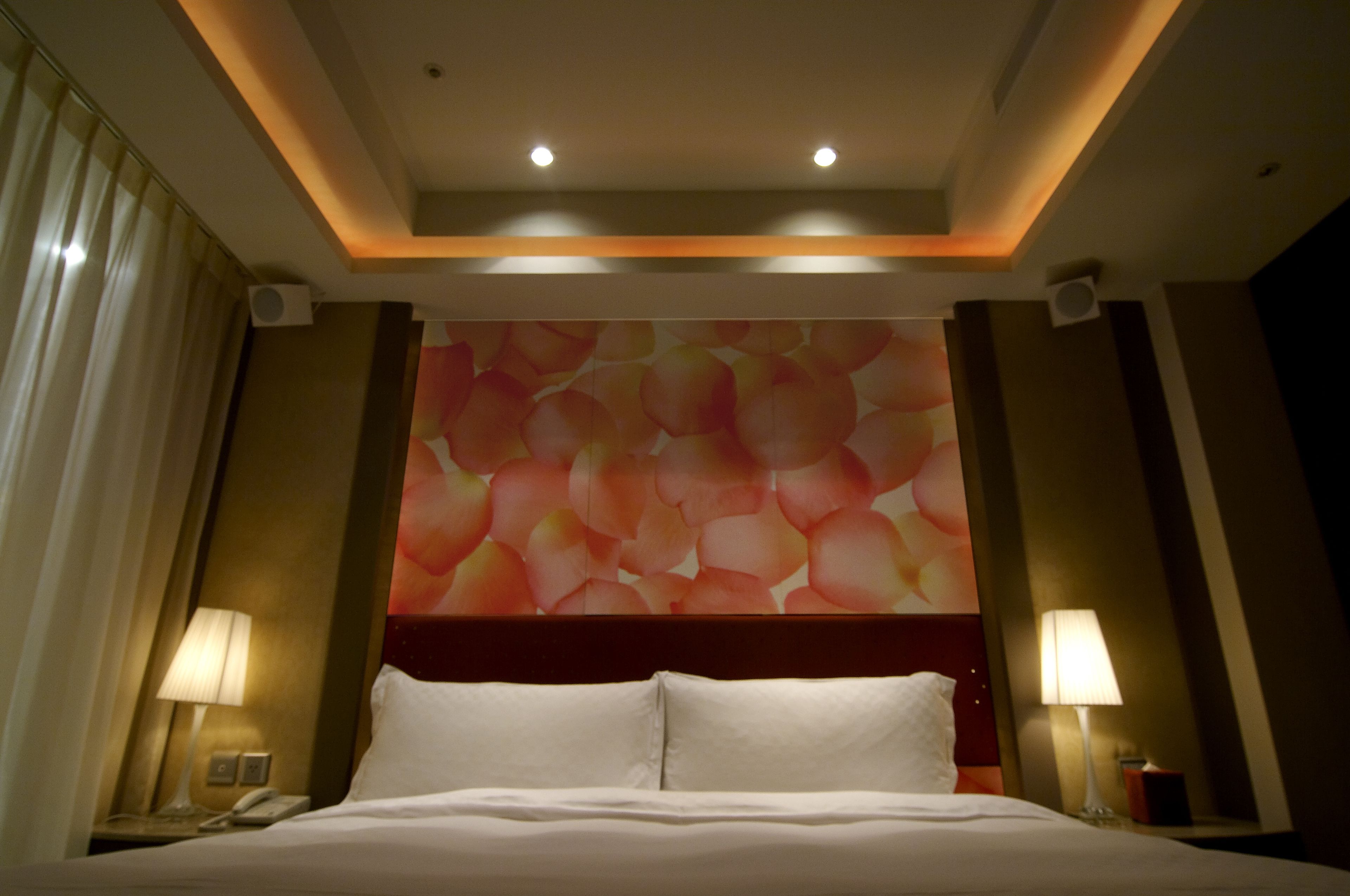 A hotel room with a bed, two bedside lamps, a wall with a floral pattern, and a ceiling with recessed lighting.