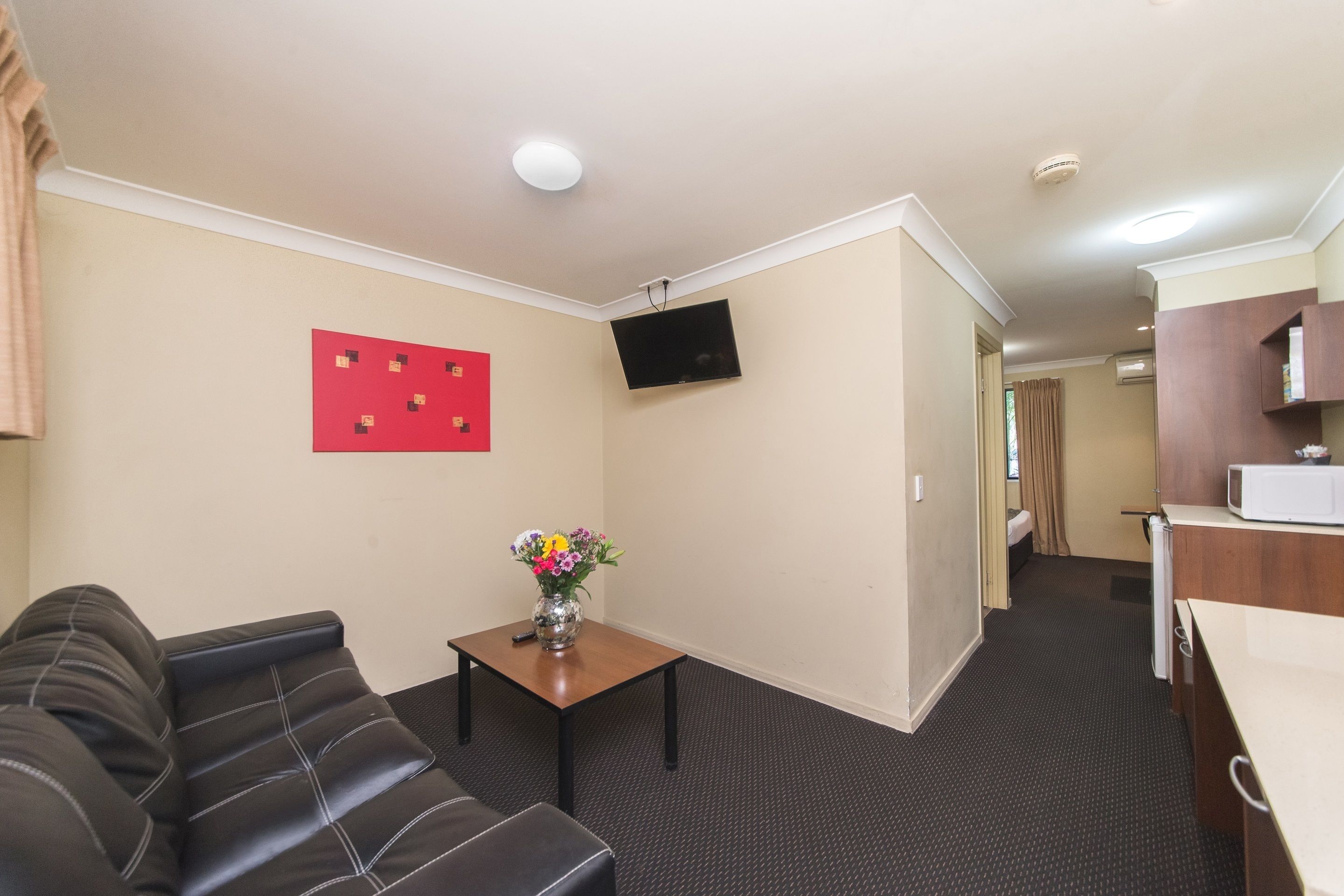 A hotel room with a sofa, a small table with a vase of flowers, a flat-screen TV mounted on the wall, a microwave, and a kitchenette area.