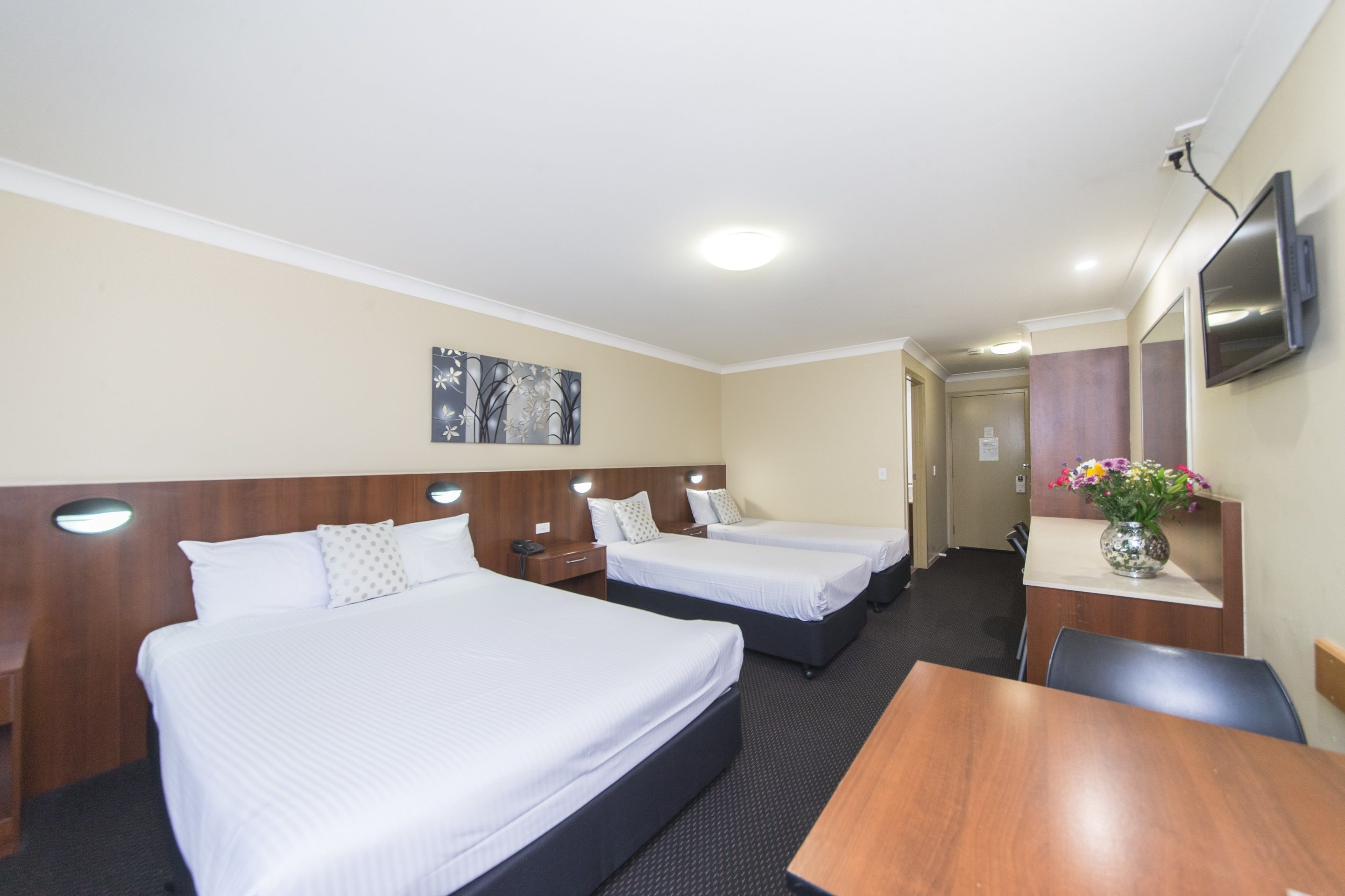 A hotel room with three single beds, a wooden desk, a TV mounted on the wall, and a vase with flowers on the desk.