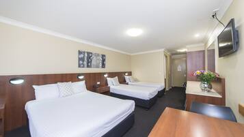 A hotel room with three single beds, a wooden desk, a TV mounted on the wall, and a vase with flowers on the desk.