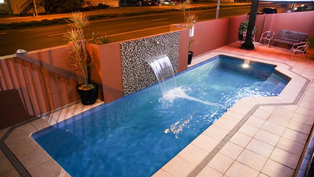 Piscina retangular ao ar livre com cachoeira, rodeada por um deck ladrilhado e vasos de plantas.