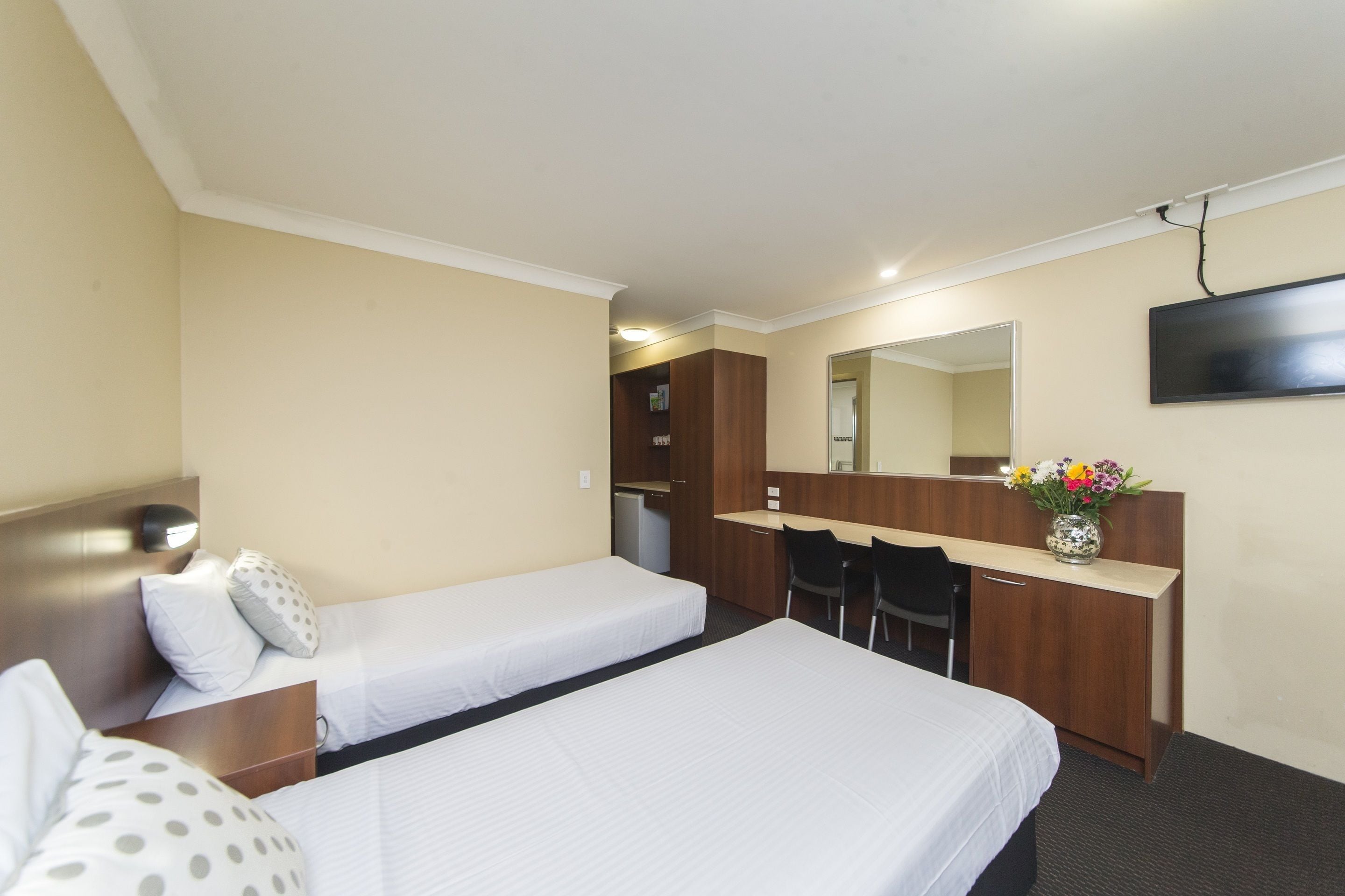 A hotel room with two single beds, a desk with a chair, a mirror, and a TV mounted on the wall.