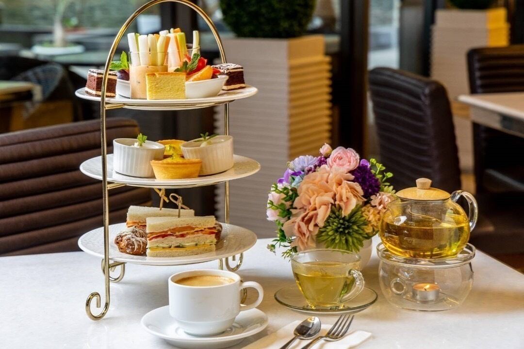 A three-tiered dessert stand with various pastries and sandwiches, a cup of coffee, a teapot with tea, and a floral arrangement on a table.