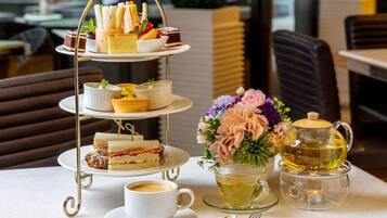 A three-tiered dessert stand with various pastries and sandwiches, a cup of coffee, a teapot with tea, and a floral arrangement on a table.