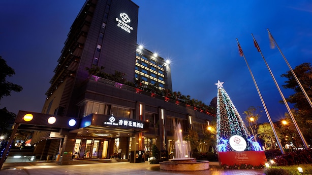 Un hotel de diseño moderno, con un logo destacado en la fachada, un gran árbol de Navidad iluminado y una fuente en el patio.