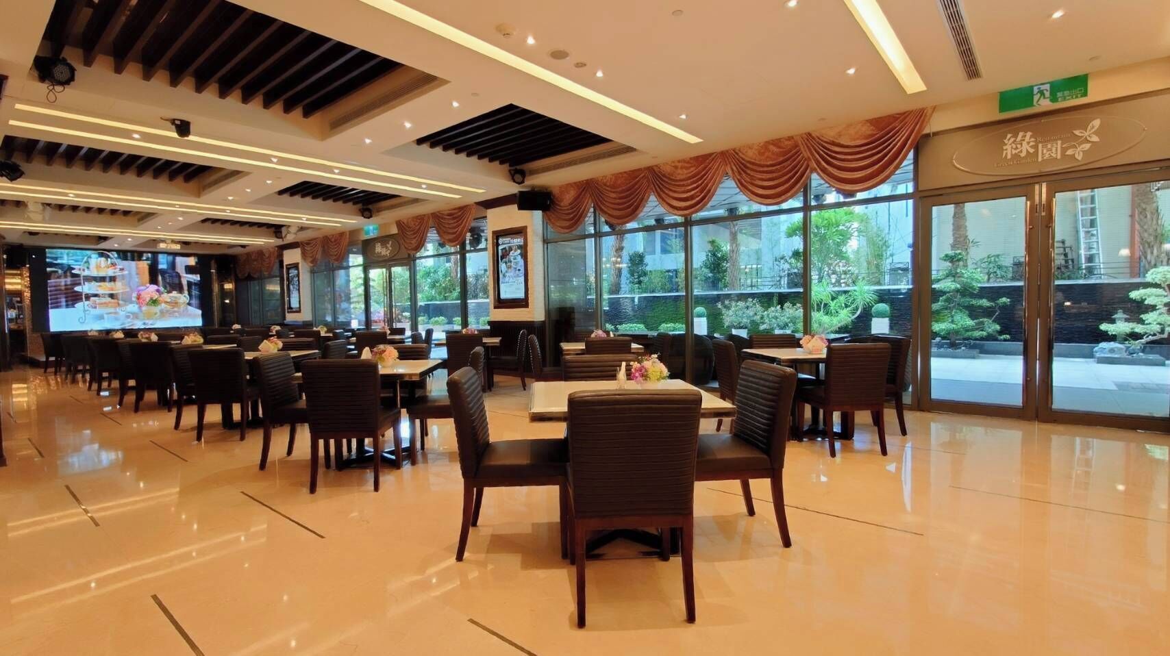 A spacious restaurant with multiple tables and chairs, large windows offering a view of the outdoors, and a modern ceiling design with recessed lighting.