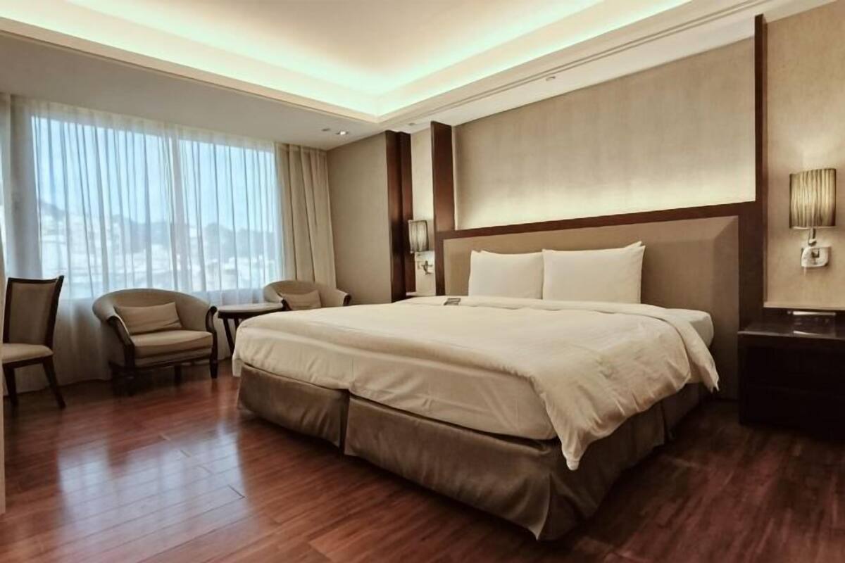 Ein Hotelzimmer mit einem großen Bett, einem Schreibtisch, einem Sessel und Blick auf die Stadt durch das Fenster.