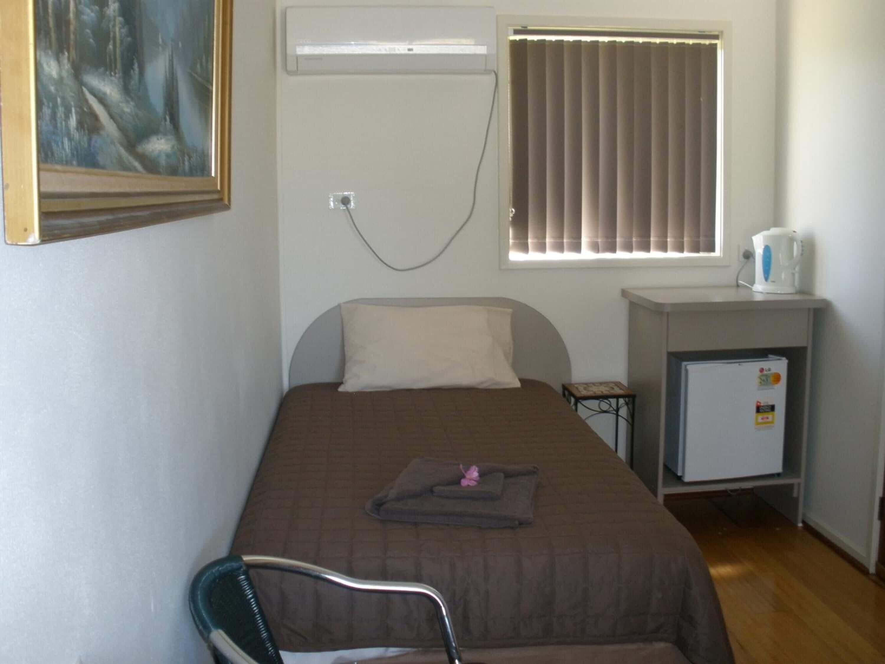 Una habitación pequeña con cama individual, una cama, una silla, un escritorio con refrigerador y una ventana con persianas.