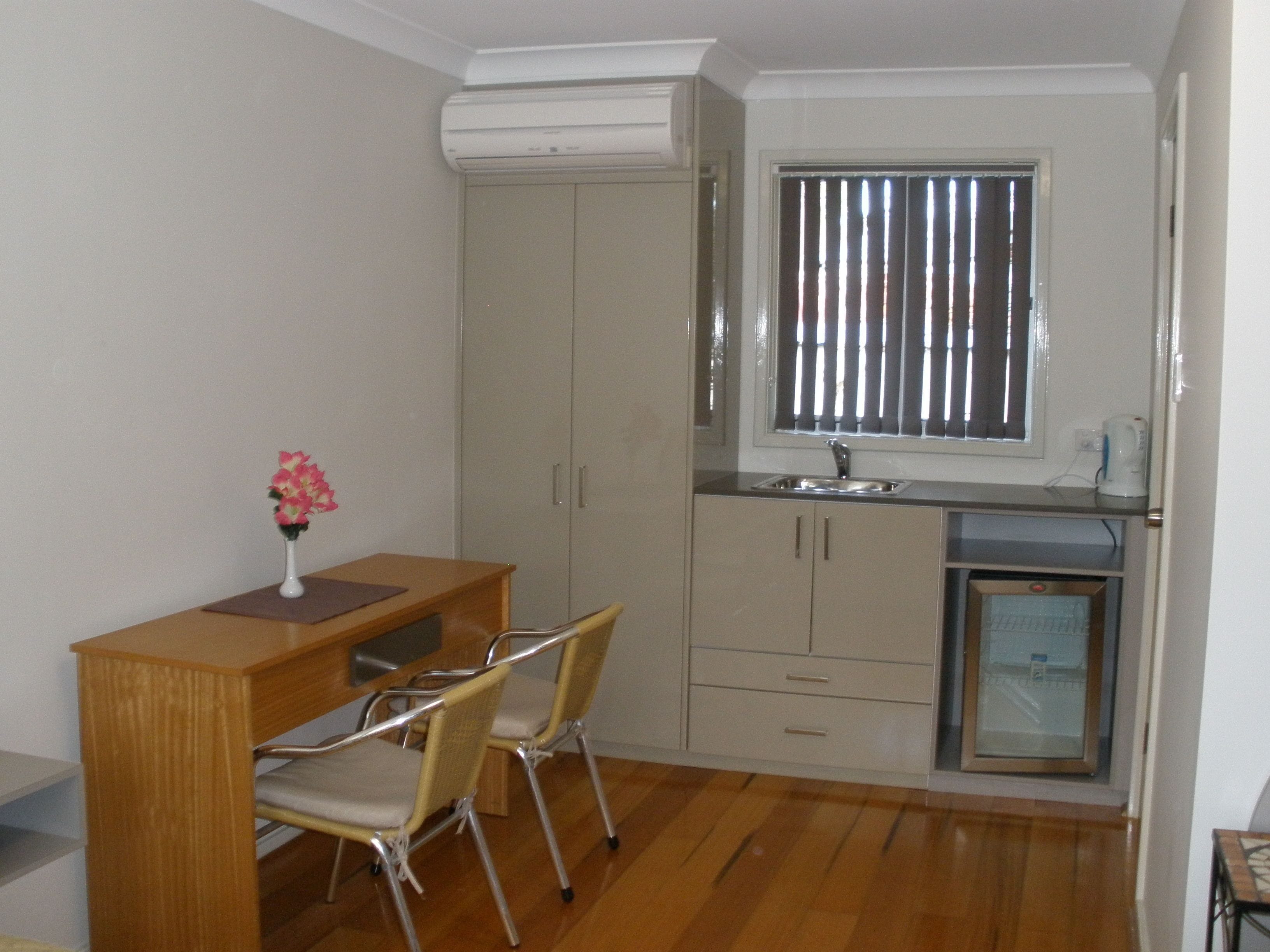 A compact kitchen with a wooden table, two chairs, a small refrigerator, and a window with blinds.