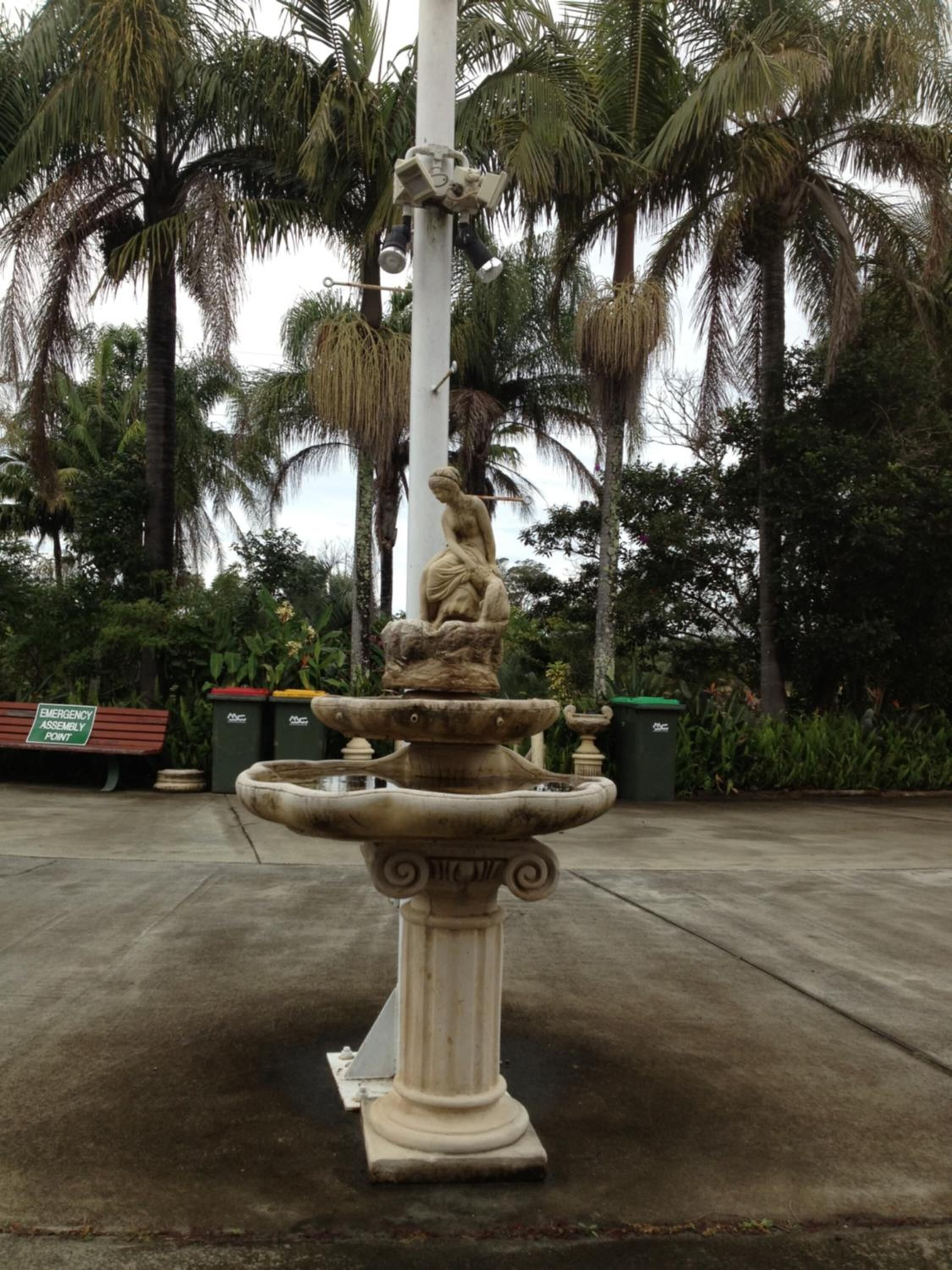 Une fontaine ornée d’une statue représentant une femme tenant un enfant, le tout étant entouré de palmiers.