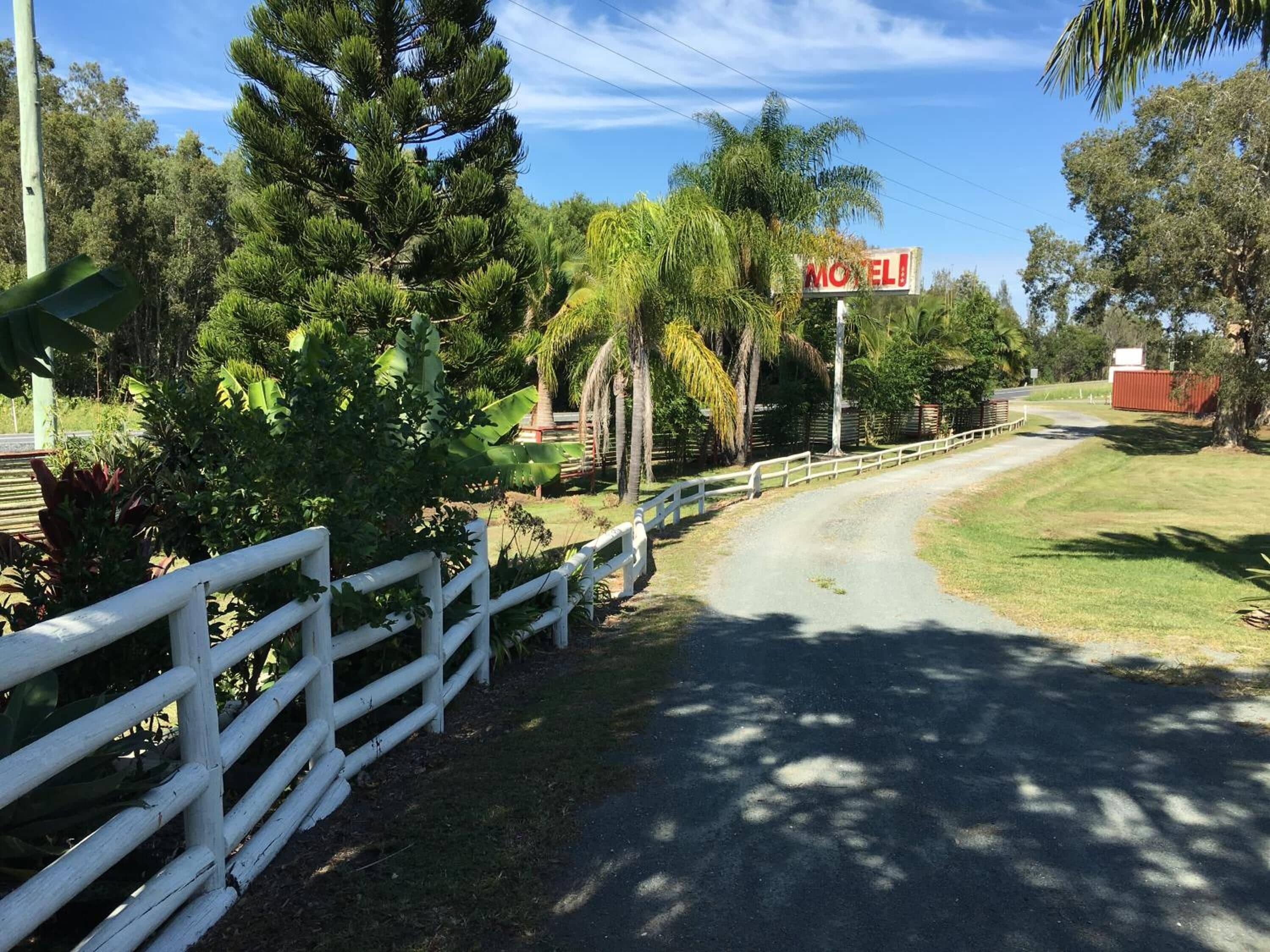 A rural road leading to a motel with a white fence and lush greenery.