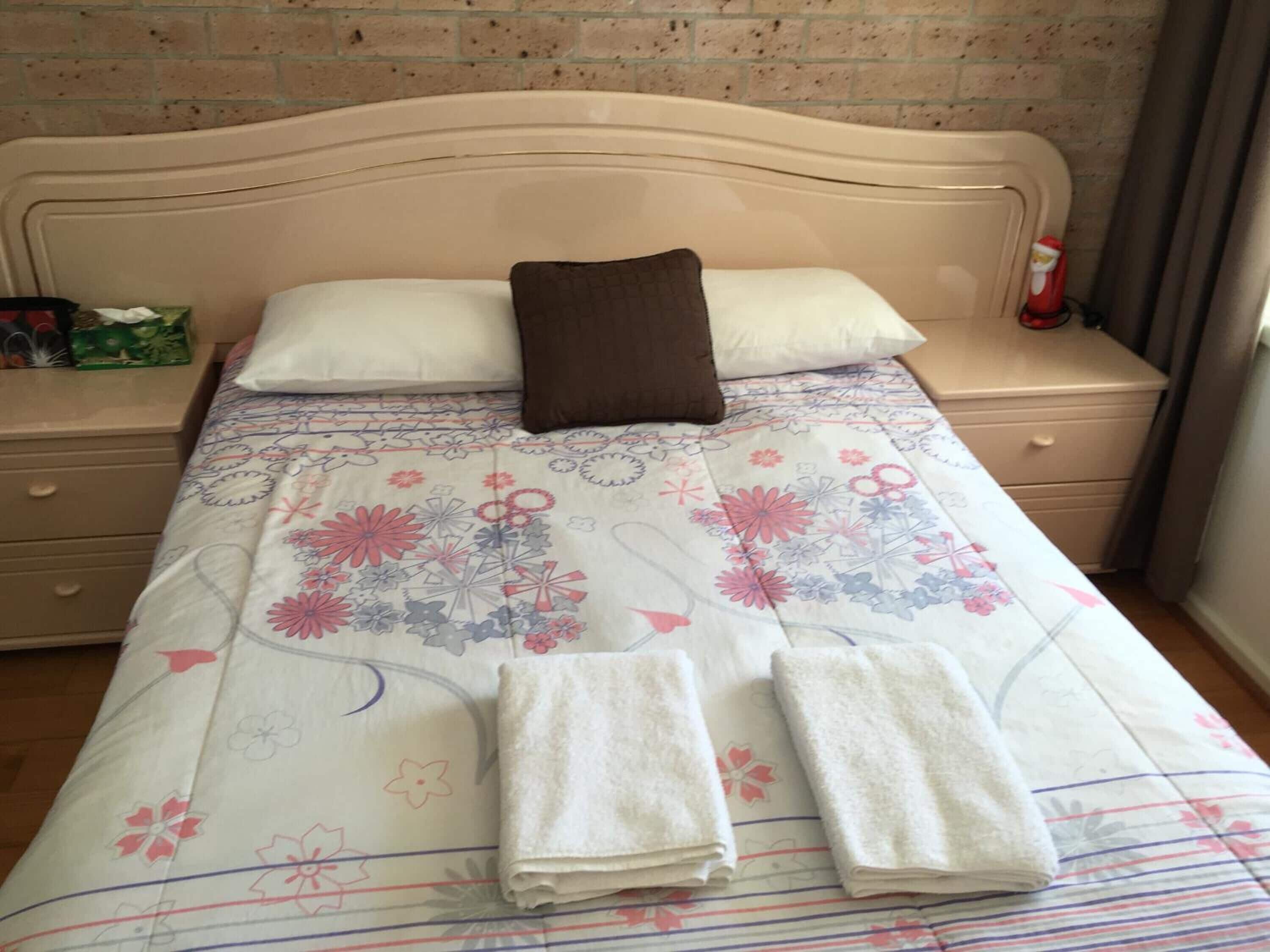 Cama bem arrumada com roupa de cama floral, dois travesseiros e duas toalhas dobradas.