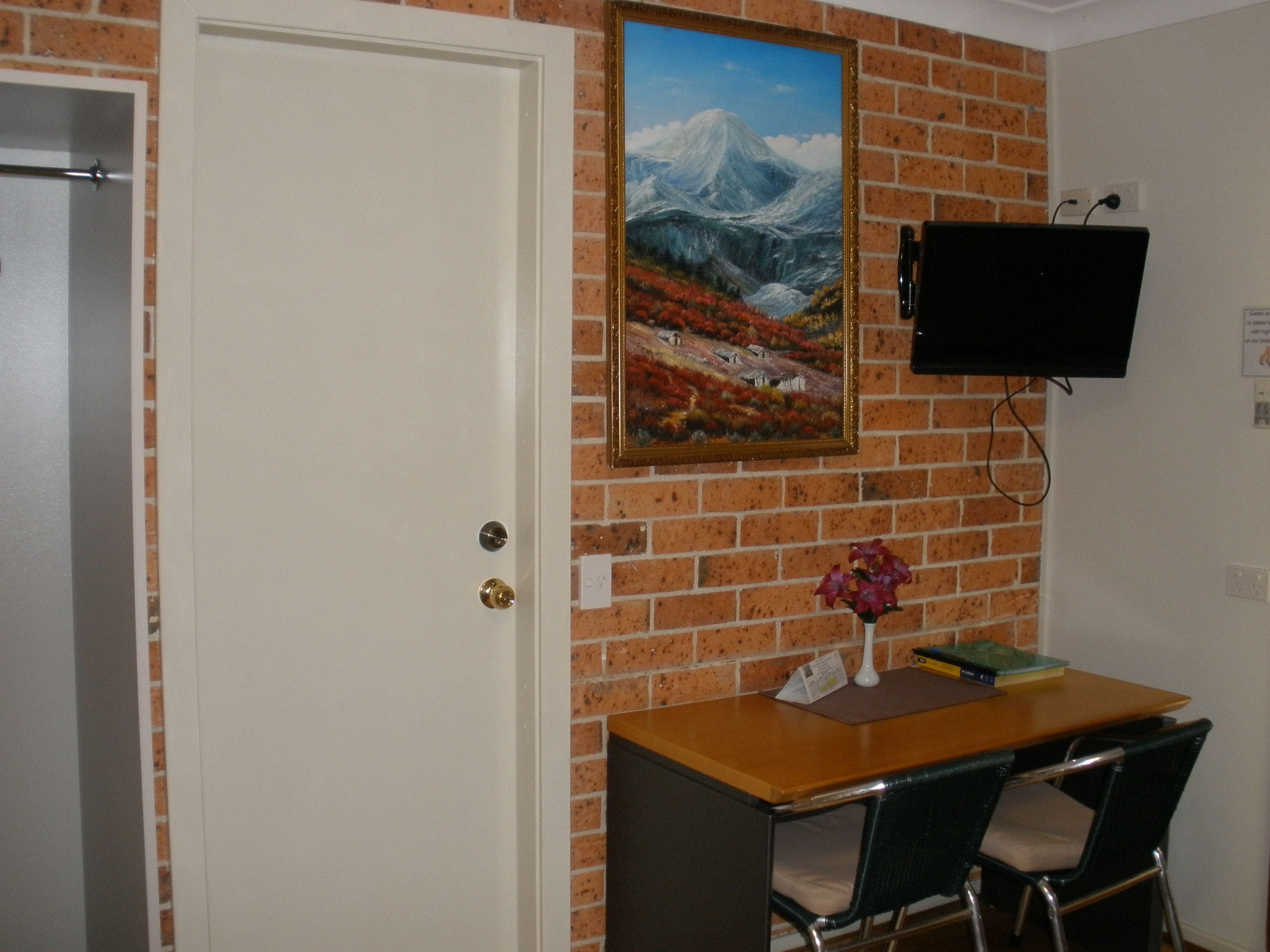 A room with a brick wall, a white door, a painting of a mountain landscape, a flat-screen TV mounted on the wall, a wooden desk with two chairs, and a vase with flowers on the desk.