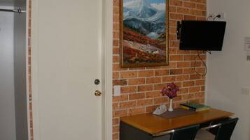 A room with a brick wall, a white door, a painting of a mountain landscape, a flat-screen TV mounted on the wall, a wooden desk with two chairs, and a vase with flowers on the desk.