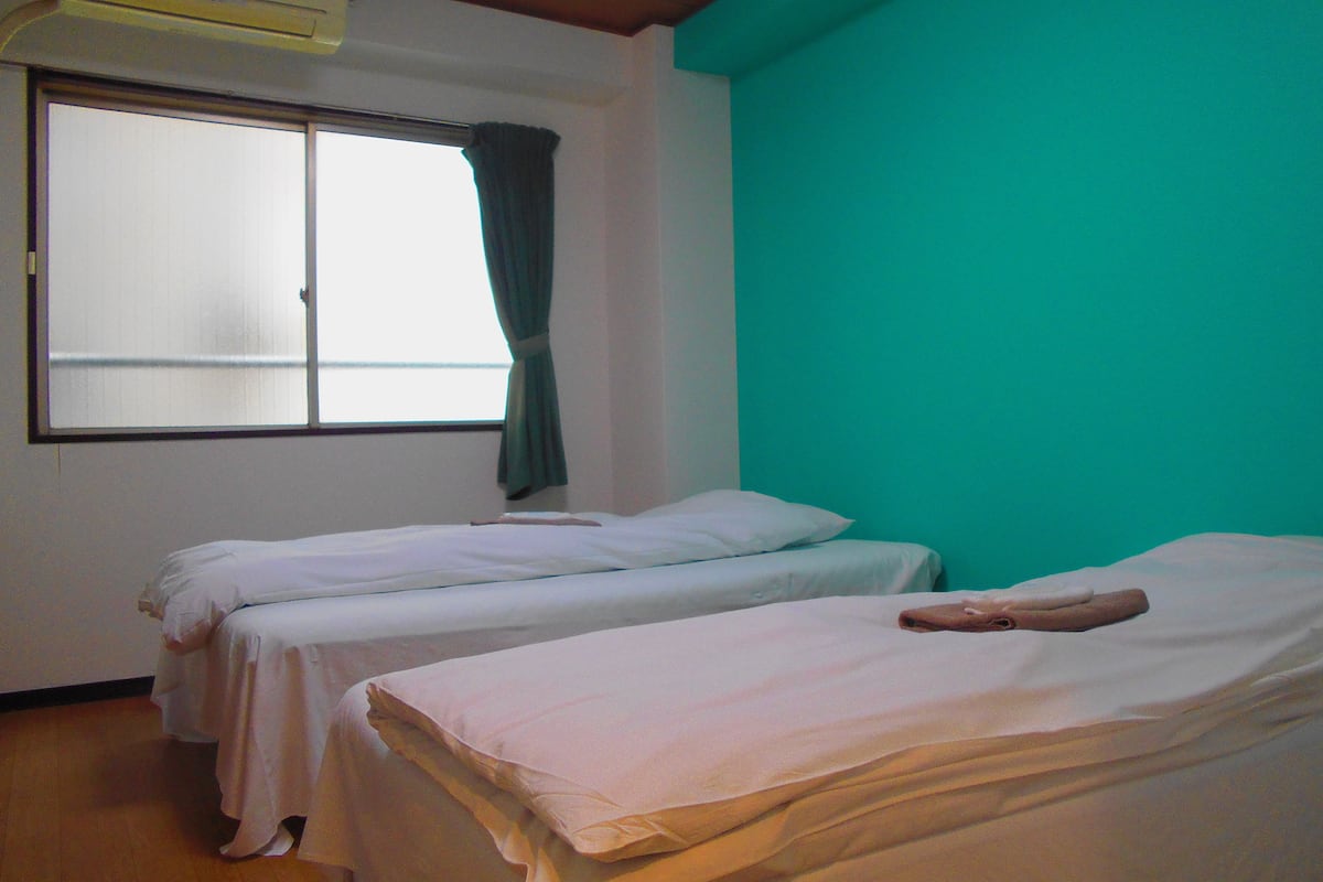 A room with two single beds, a window with curtains, and a teal-colored wall.