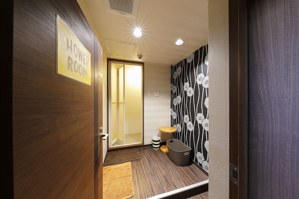 Une chambre d’hôtel avec une porte en bois, un panneau indiquant la présence d’une salle de douche, une petite table ronde, une poubelle et un mur décoré d’un motif.