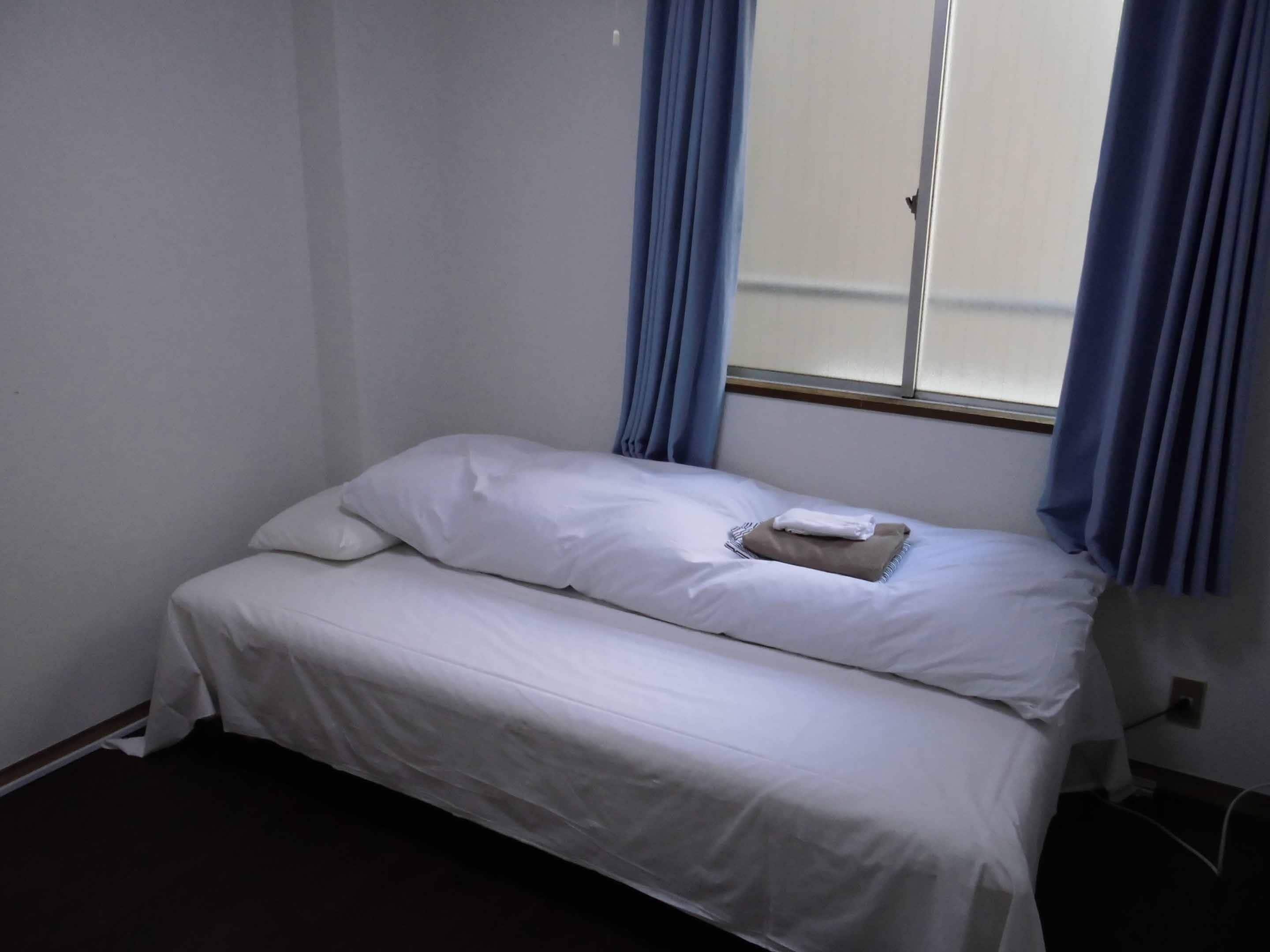Une petite chambre avec un lit simple, du linge de lit blanc, une serviette pliée sur le lit et des rideaux bleus aux fenêtres.