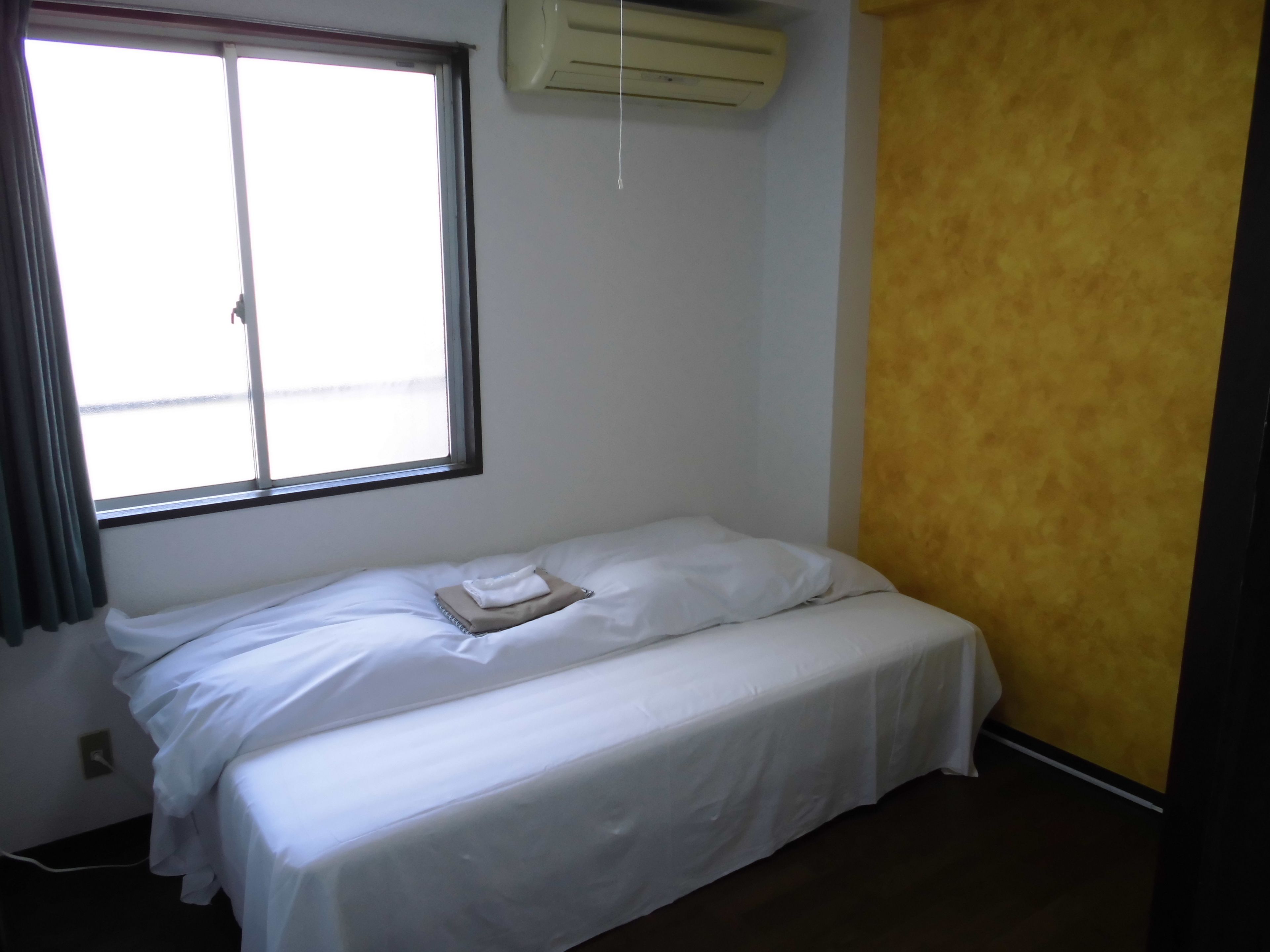Une petite chambre avec un lit simple, une fenêtre, un appareil de climatisation et un mur d’accent jaune.