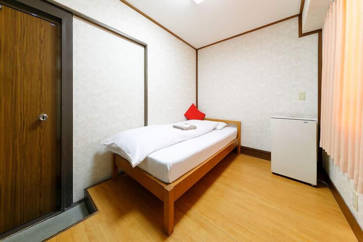 A small, neatly arranged bedroom with a bed, a red pillow, a white bedside table, and a white refrigerator.