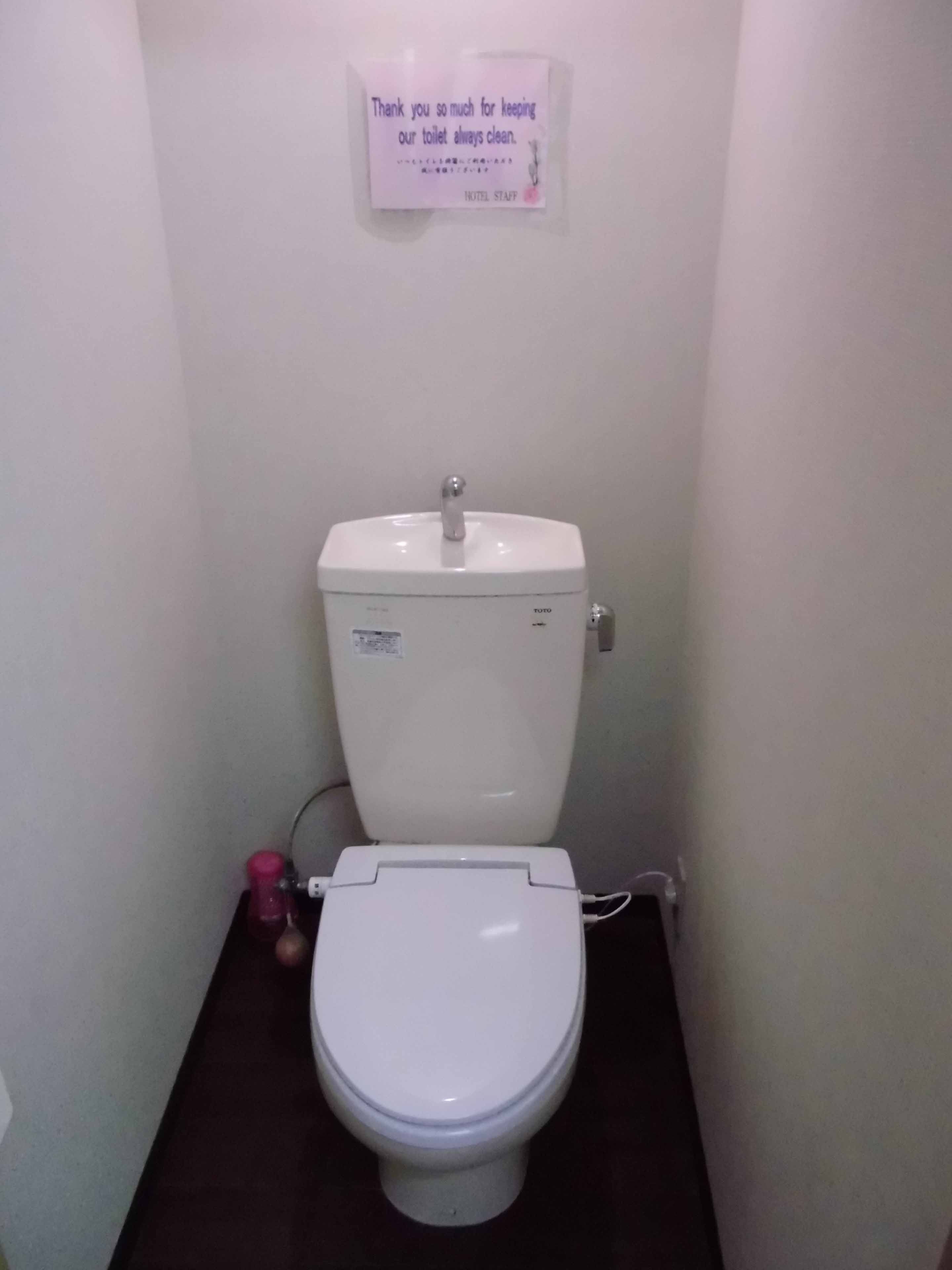 A white toilet with a TOTO brand label, a sign above thanking for keeping the toilet clean, and a pink bottle on the floor.