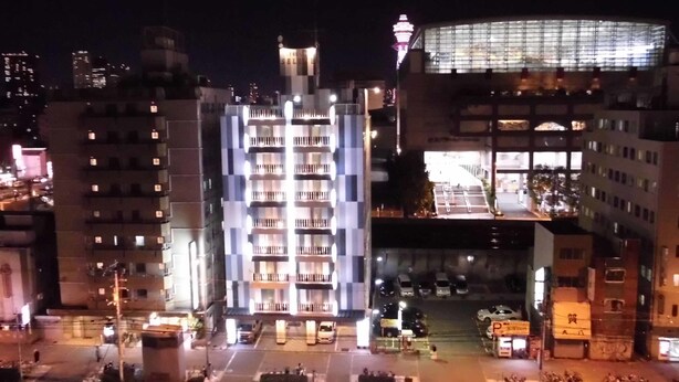 A multi-story building with balconies, a parking lot, and a brightly lit tower in the background. - Hotel Taiyo (Osaka)