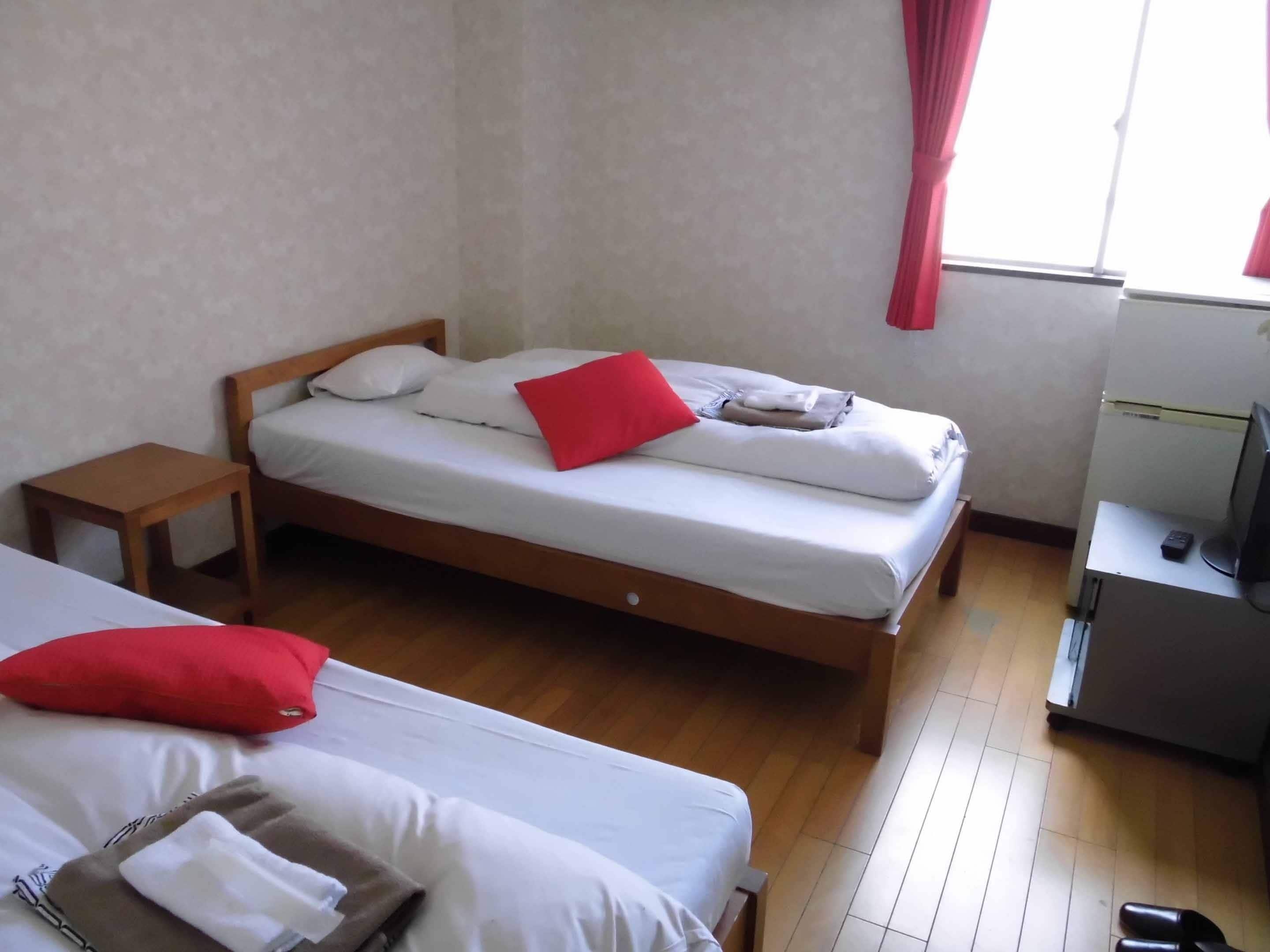 Ein Zimmer mit zwei Betten, Holzboden, einem kleinen Tisch, einem Fernseher und einem Fenster mit Vorhängen.