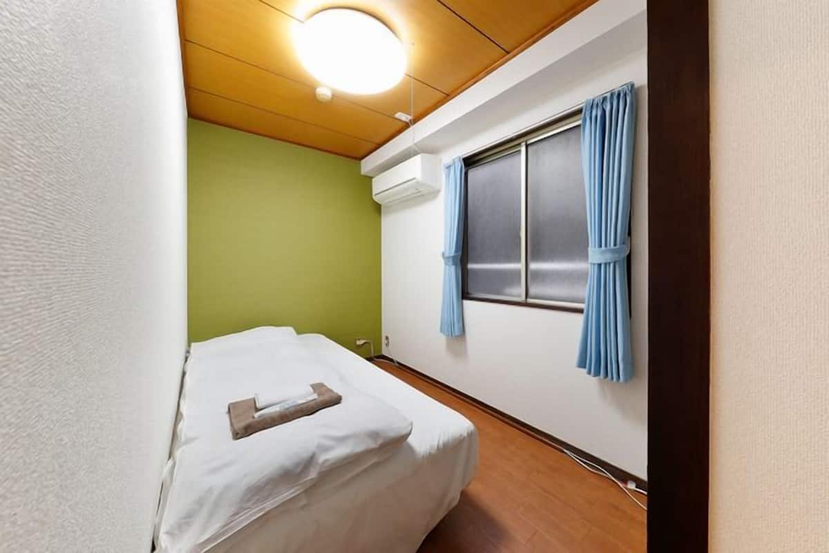 A small, narrow hotel room with a single bed, a window with curtains, a wooden ceiling, and a wall-mounted air conditioner.