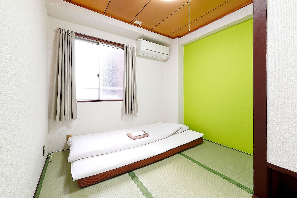 A small room with a bed, a window with curtains, an air conditioning unit, and a green wall.