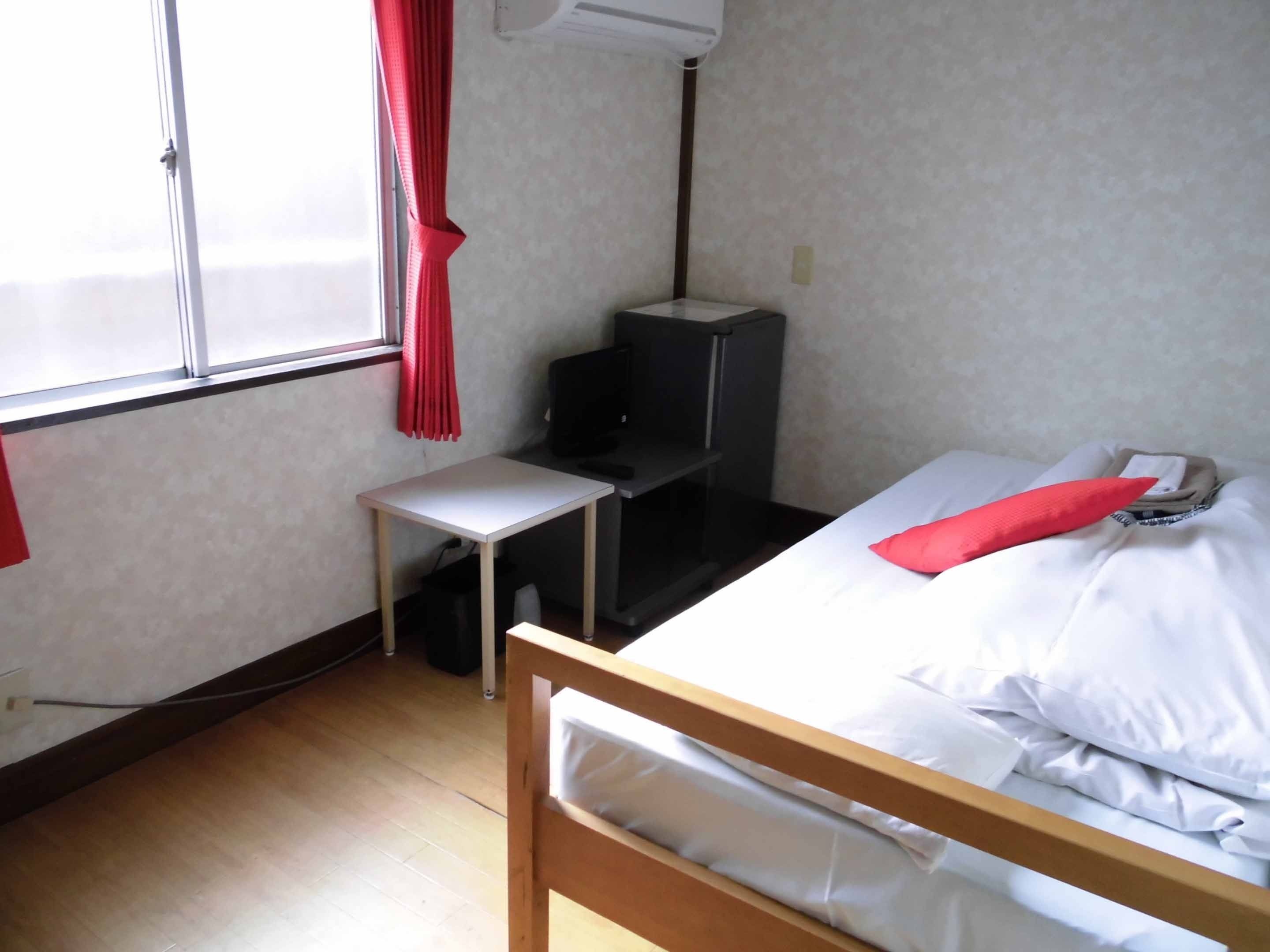 A small, simple room with a bed, a desk, and a window with red curtains.