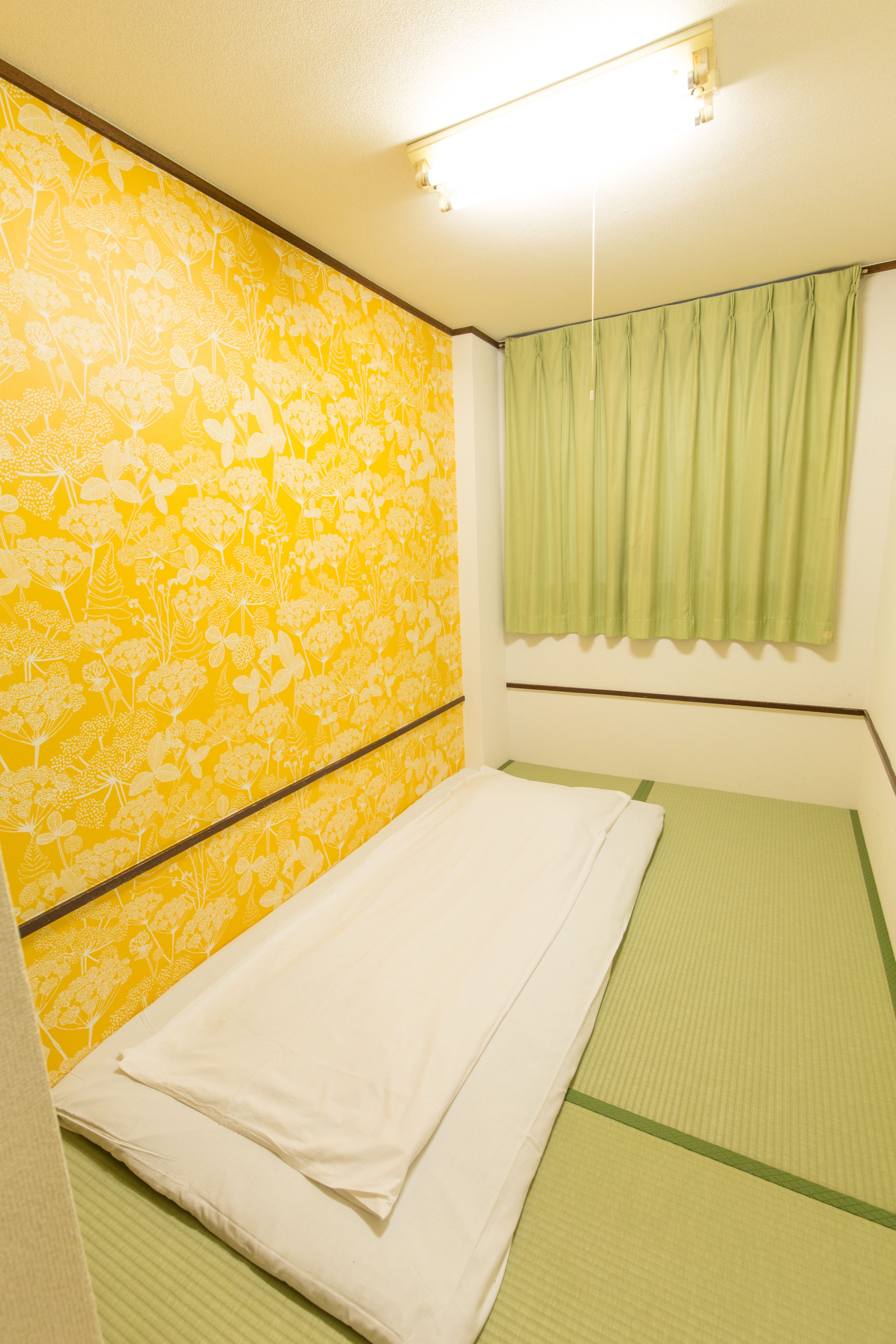 A room with a bed, green tatami flooring, a yellow floral wallpaper, and a green curtain.