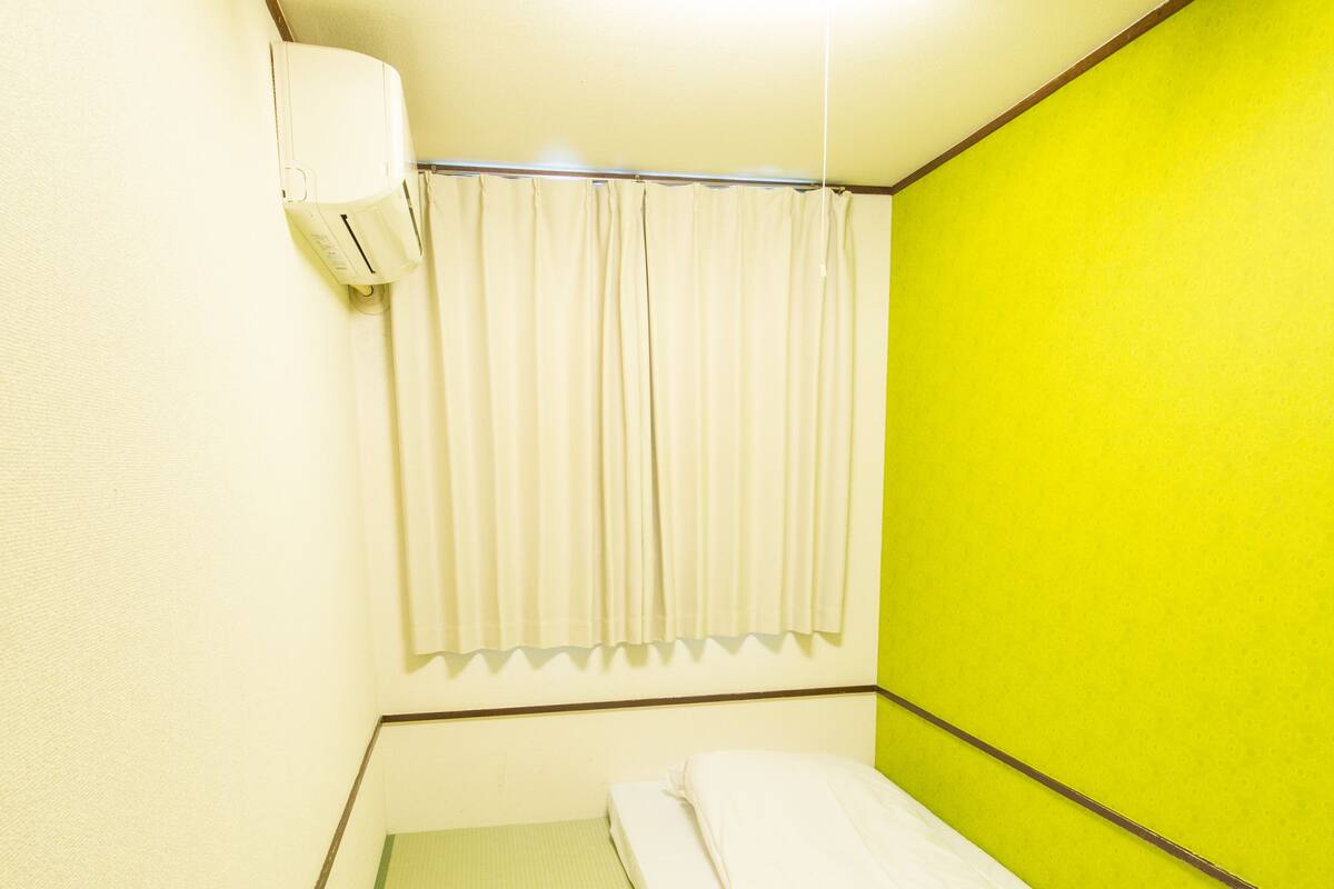 Une petite chambre avec un lit simple, un mur d’accent vert, un climatiseur blanc et une fenêtre avec des rideaux blancs.