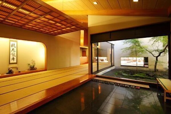 Un interior tradicional japonés con un mostrador de recepción de madera, un ventanal con vistas a un árbol y un edificio, y un suelo de baldosas.