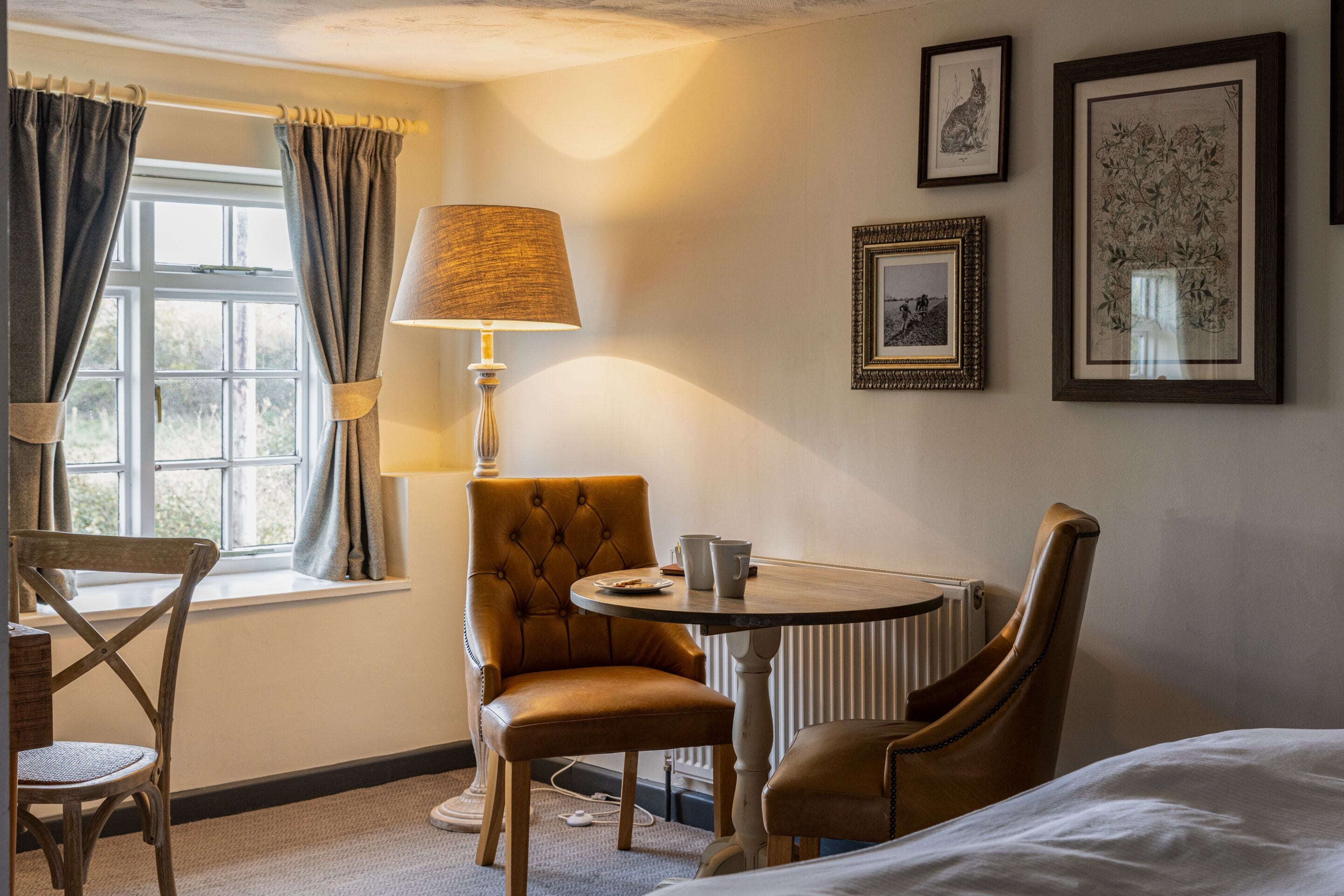 Una habitación de hotel con una cama, una mesa con lámpara, una silla, una ventana con cortinas y cuadros en la pared.