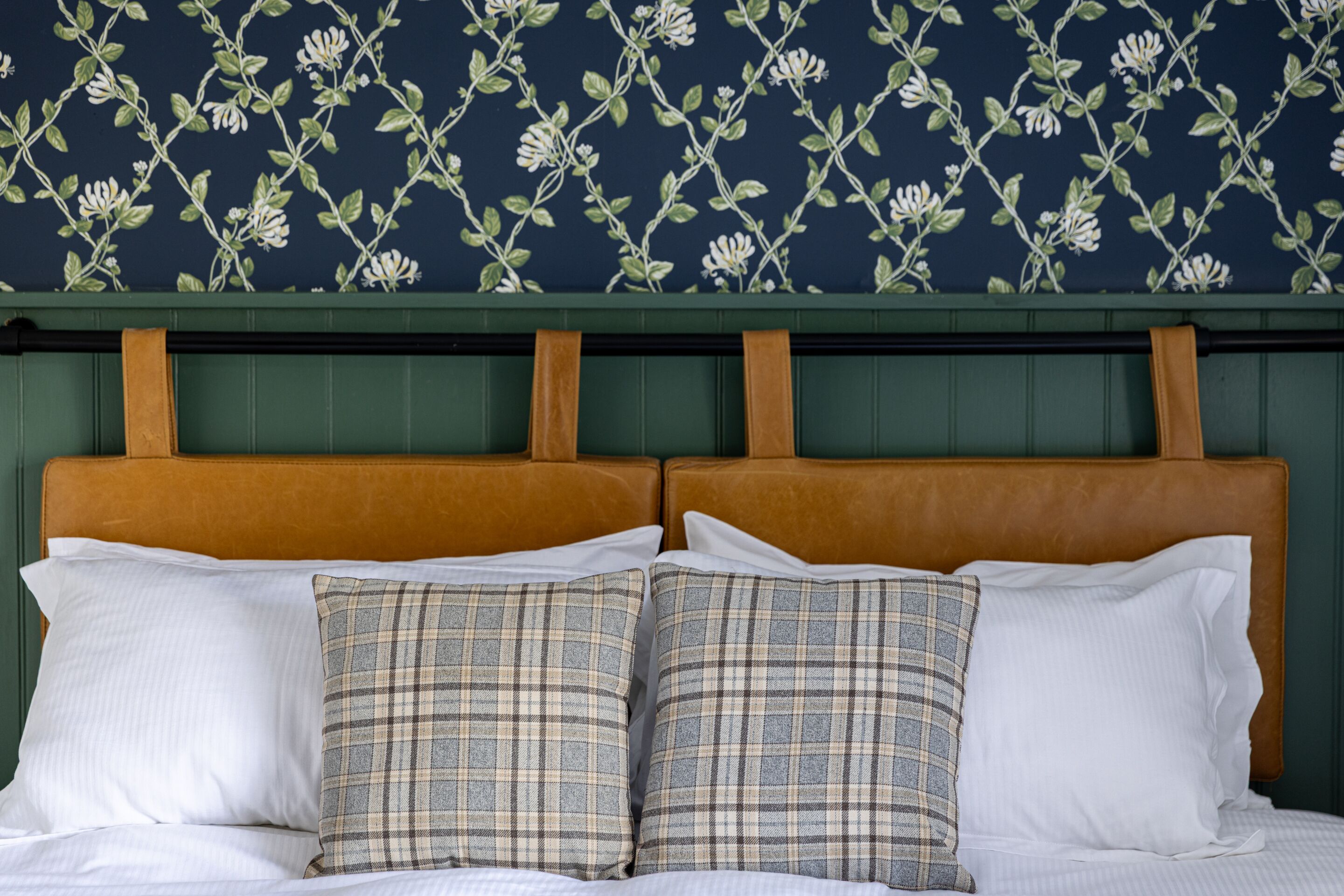 Una cama con almohadas blancas y a cuadros, un cabecero de madera y un fondo de papel tapiz con motivos florales.