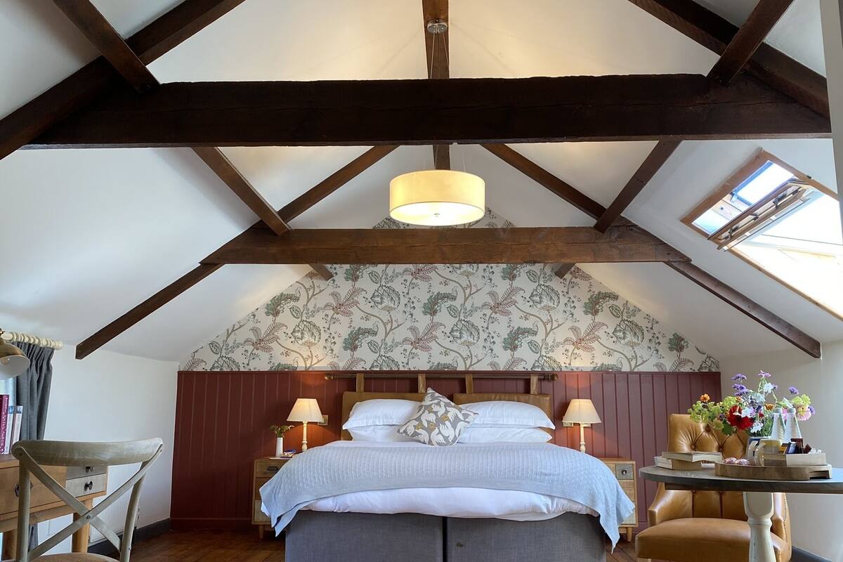 A bedroom with a sloped ceiling, a bed with a headboard, two bedside tables with lamps, a chair, and a small table with flowers.