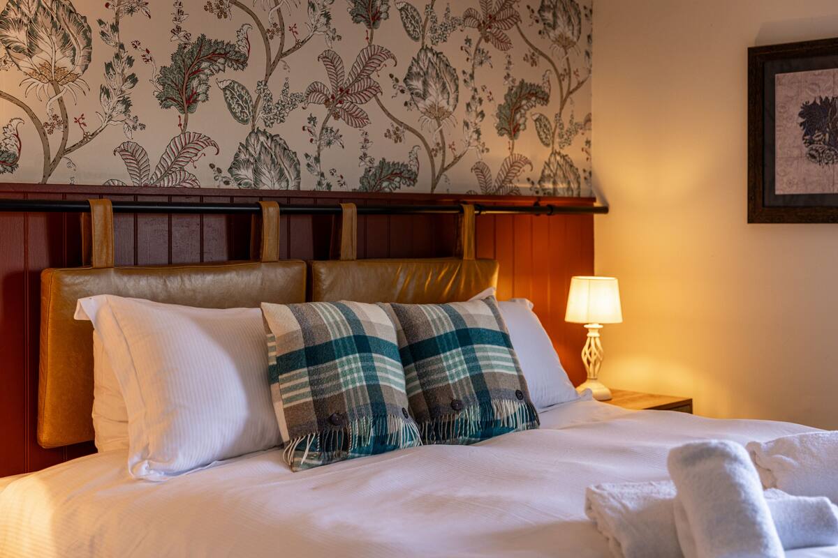 A neatly made bed with white linens, two plaid throw pillows, and a headboard with a floral wallpaper design.