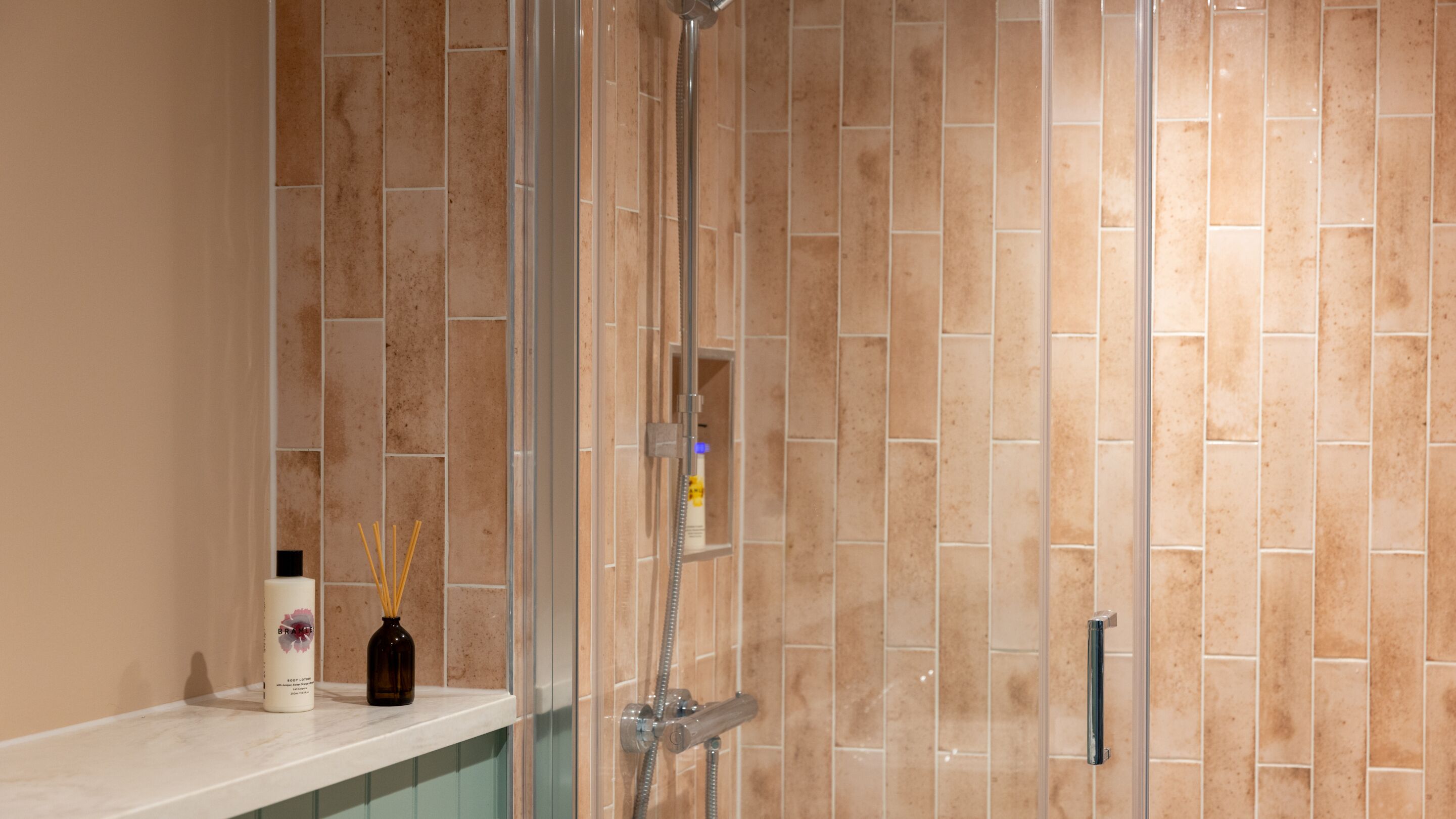 A bathroom with a glass-enclosed shower, beige tiled walls, a marble countertop, and a bottle of lotion.