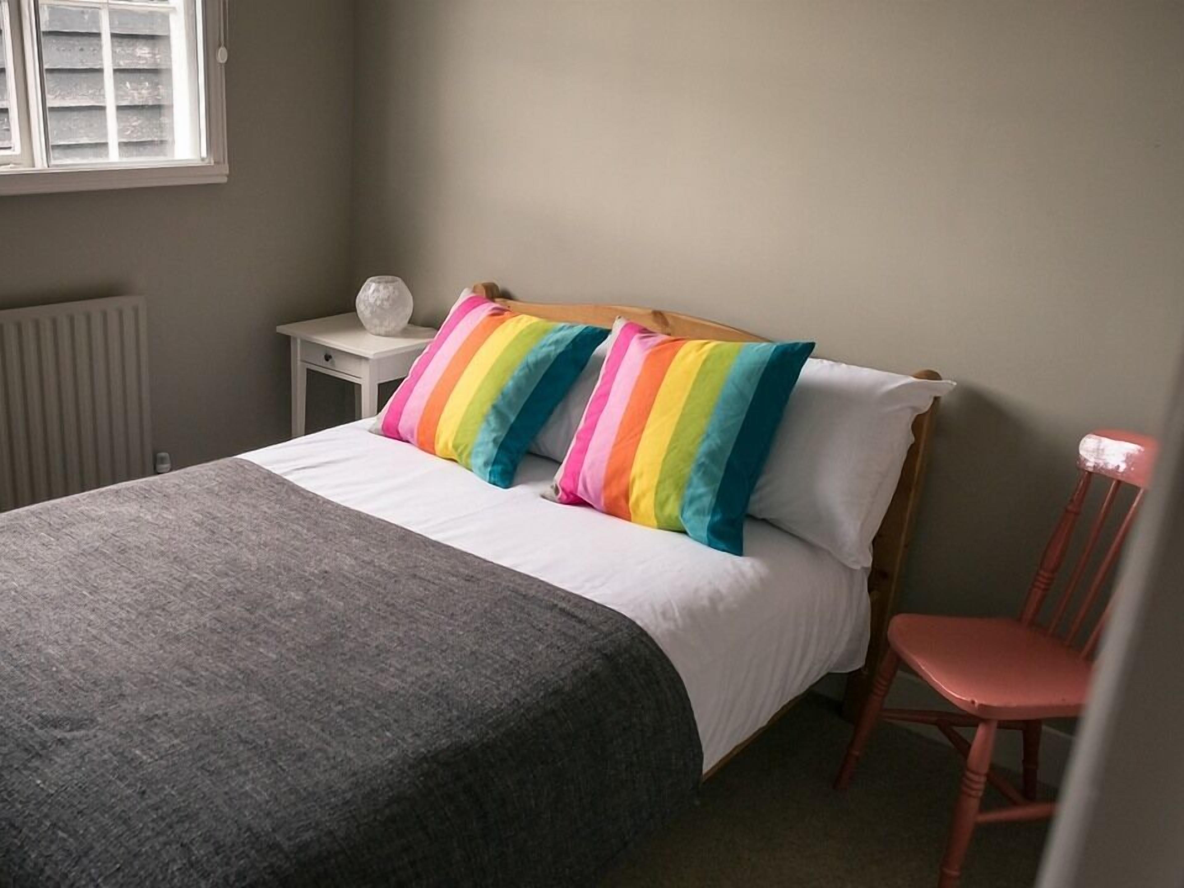 A neatly made bed with a grey blanket, a white pillow, and two rainbow-striped pillows. A small white nightstand with a white object on it is beside the bed. A red chair is positioned in front of the bed.