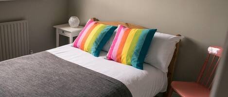 A neatly made bed with a grey blanket, a white pillow, and two rainbow-striped pillows. A small white nightstand with a white object on it is beside the bed. A red chair is positioned in front of the bed.