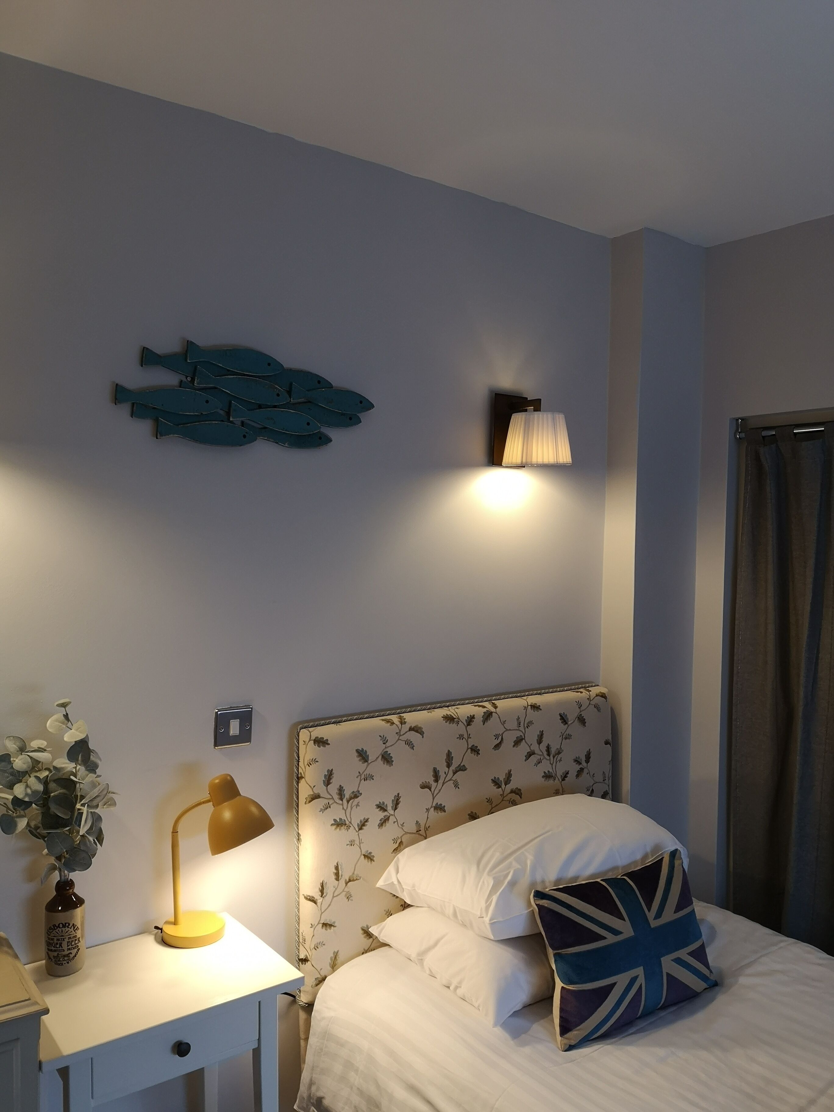 A bedroom with a bed, a nightstand, a lamp, a wall-mounted light, and a decorative fish wall art piece.