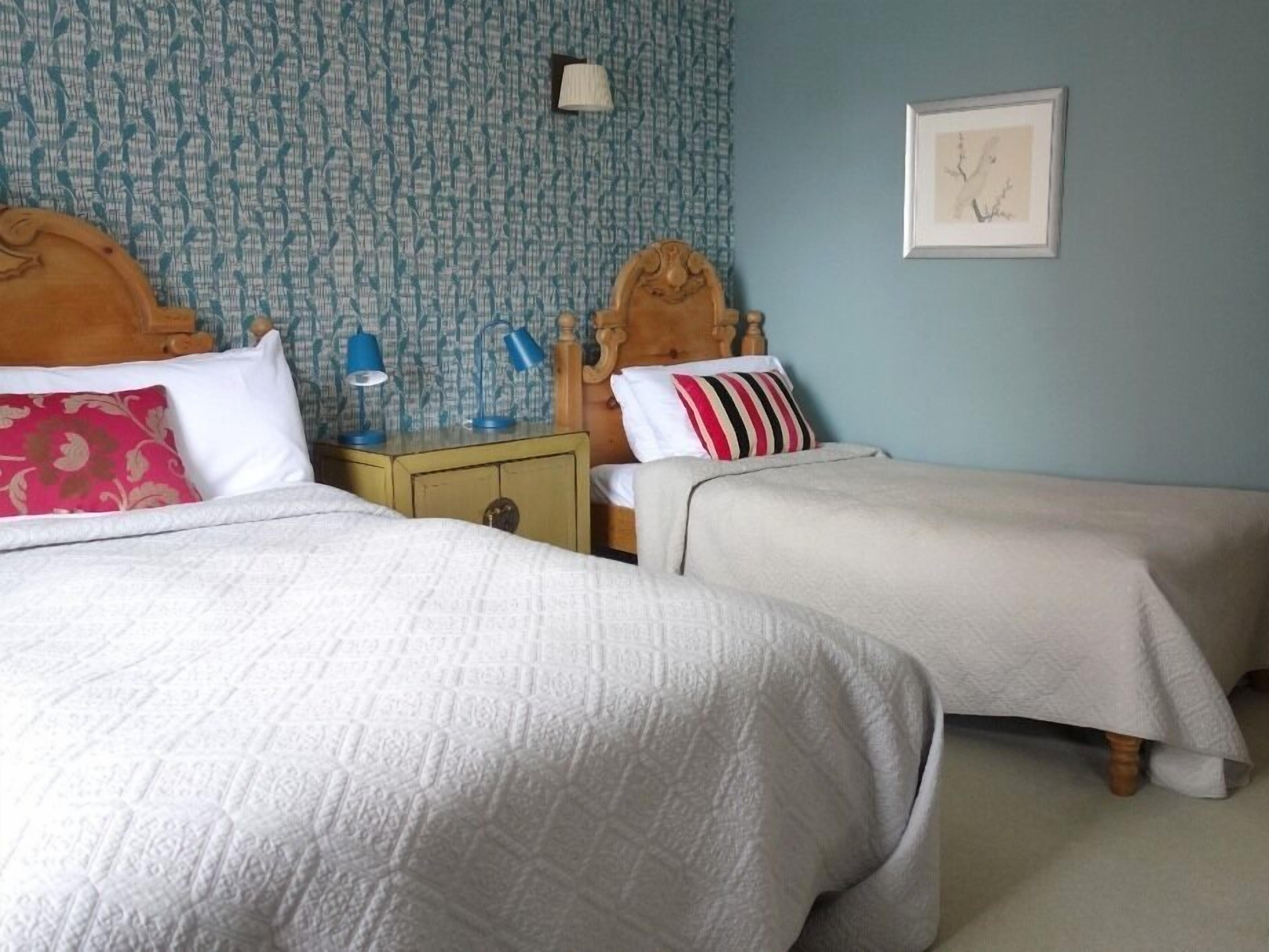 A room with two beds, a nightstand with a lamp, and a wall with a patterned wallpaper and a framed picture.