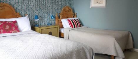 A room with two beds, each with a wooden headboard, a nightstand with a lamp, and a wall with a patterned wallpaper and a framed picture.