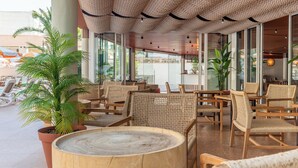 Un amplio espacio al aire libre con mobiliario de mimbre y una mesa redonda de madera, rodeado de ventanas grandes y plantas en macetas.