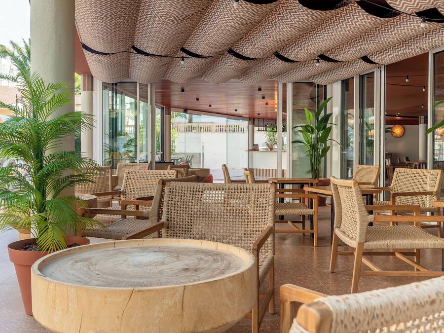 A spacious outdoor seating area with wicker furniture and a round wooden table, surrounded by large windows and potted plants.