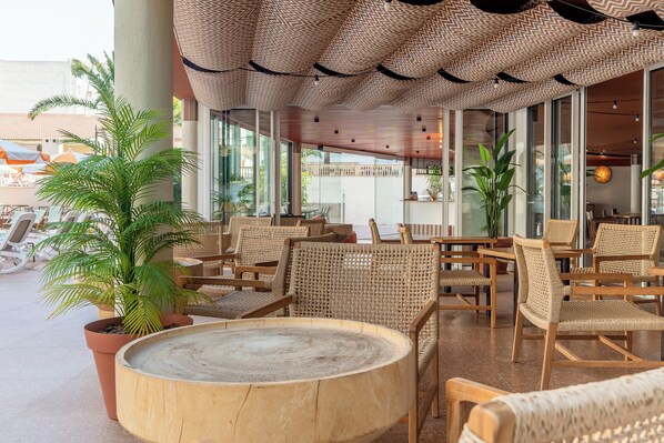 A spacious outdoor seating area with wicker furniture and a round wooden table, surrounded by large windows and potted plants.