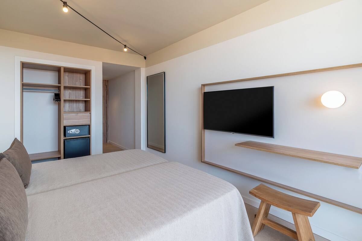 A modern hotel room with a large bed, a TV mounted on the wall, a wooden bench, and a walk-in closet.