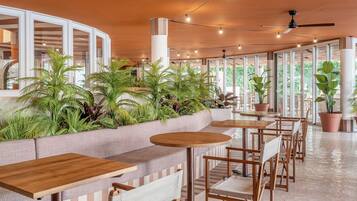 Un área de estar moderna en el interior, con mesas y sillas de madera, rodeada de amplios ventanales y plantas en macetas.
